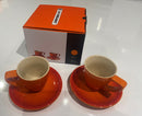 Two orange ceramic cups with saucers in front of a branded box on a white surface.